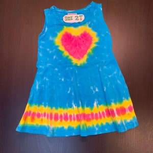 May include: A blue tie-dye sleeveless dress with a pink heart design and a yellow and pink stripe at the bottom. The dress has a tag that says "Size 2T".