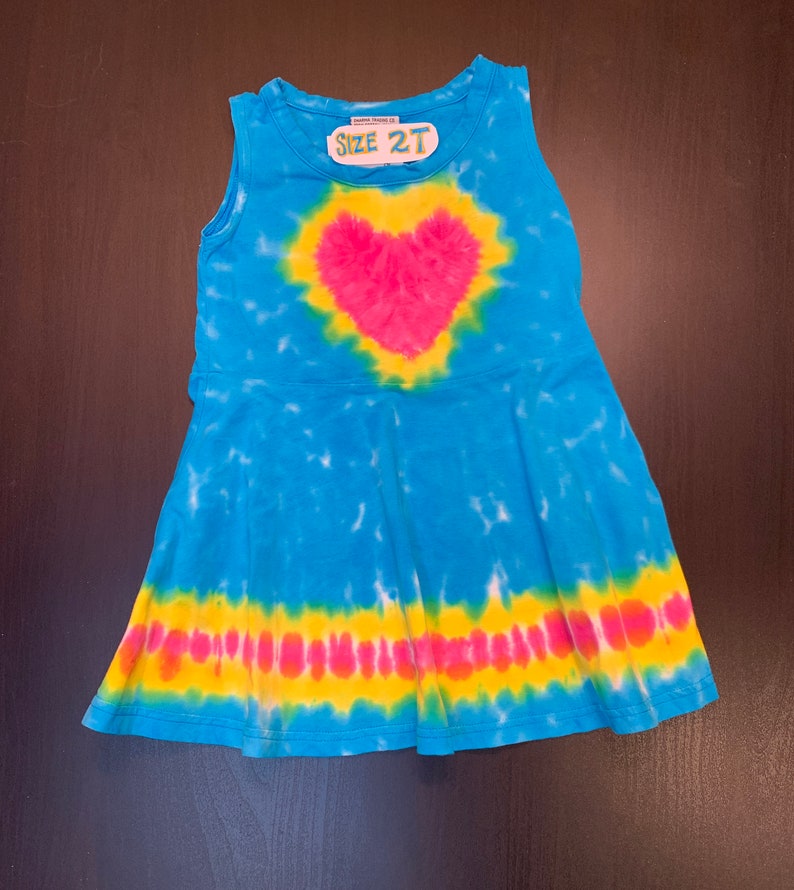 May include: A blue tie-dye sleeveless dress with a pink heart design and a yellow and pink stripe at the bottom. The dress has a tag that says "Size 2T".