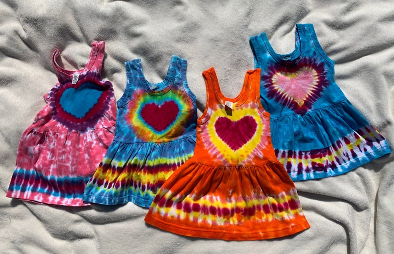 May include: Four tie-dye sleeveless dresses with a heart design in the center. The dresses are in shades of pink, blue, orange, and turquoise.