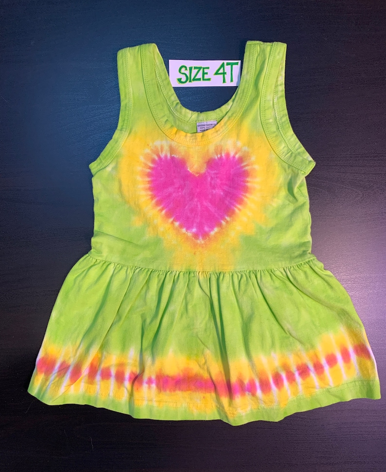 May include: Green tie-dye sleeveless dress with a pink heart design and a ruffled hem. The dress has the text "SIZE 4T" on a white tag.