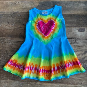 May include: A sleeveless tie-dye dress with a large heart design in shades of pink, orange, and yellow. The dress is a light blue color with a flared skirt.