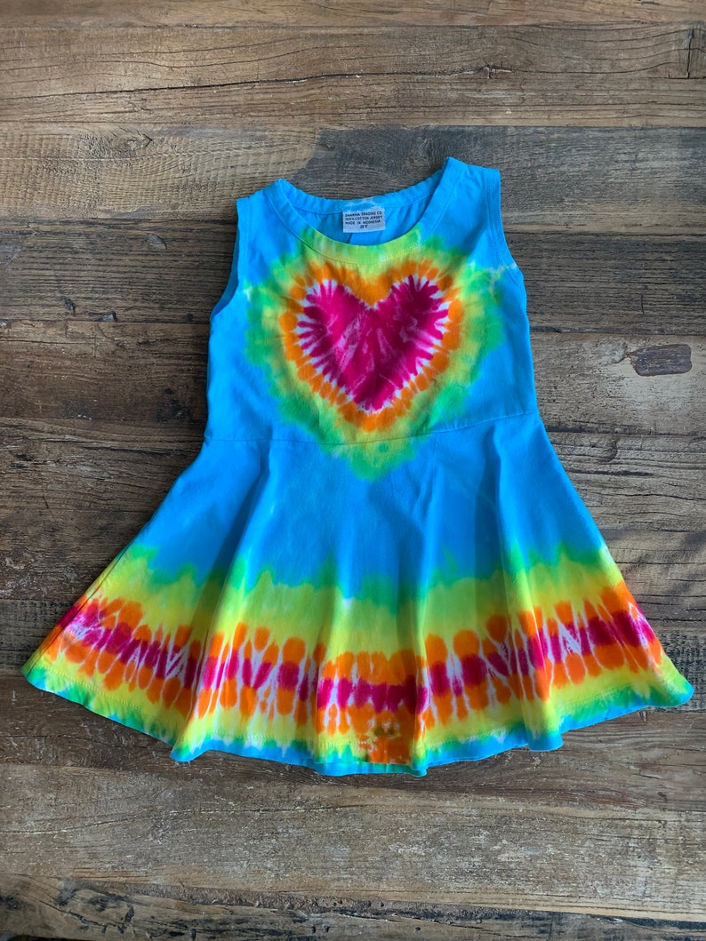 May include: A sleeveless tie-dye dress with a large heart design in shades of pink, orange, and yellow. The dress is a light blue color with a flared skirt.