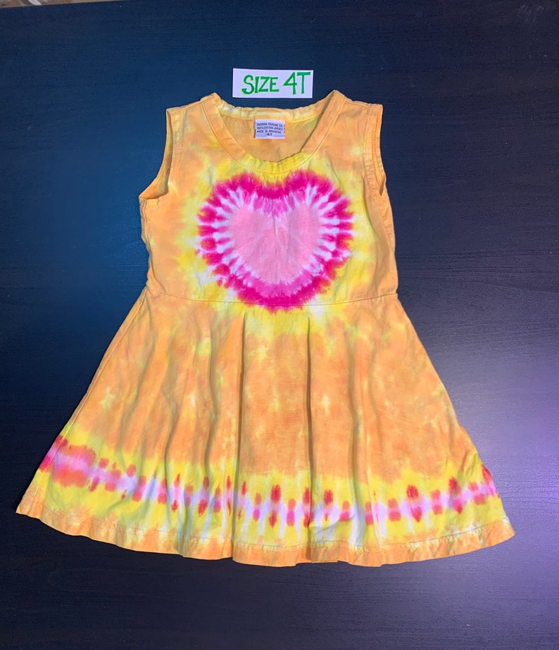 May include: A yellow tie-dye sleeveless dress with a pink heart design. The dress has a size tag that says "Size 4T".