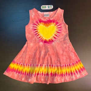 May include: A pink tie-dye sleeveless dress with a yellow heart design. The dress has a flared skirt and a size tag that says "Size 2T".