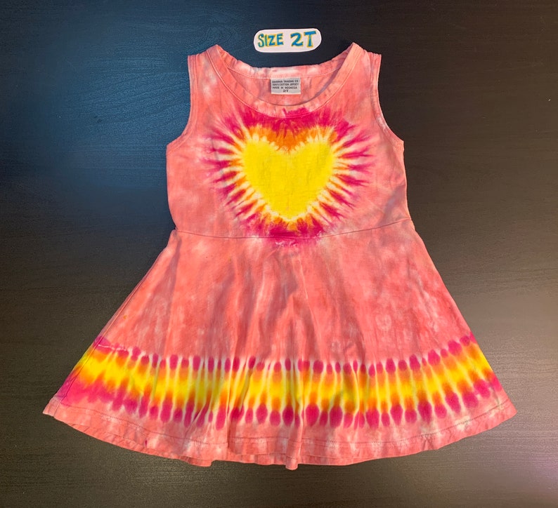 May include: A pink tie-dye sleeveless dress with a yellow heart design. The dress has a flared skirt and a size tag that says "Size 2T".