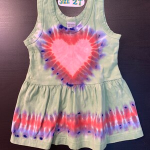 May include: Green tie-dye sleeveless dress with a pink heart design. The dress has a ruffled hem and says "Size 2T" on the front.