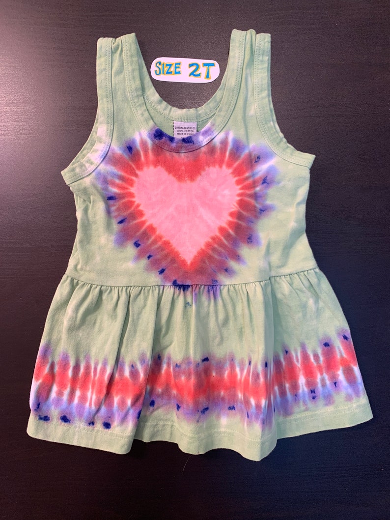 May include: Green tie-dye sleeveless dress with a pink heart design. The dress has a ruffled hem and says "Size 2T" on the front.