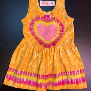 May include: A yellow, pink, and white tie-dye sleeveless dress with a heart design. The dress has a ruffled hem and says "SIZE 6" on the tag.