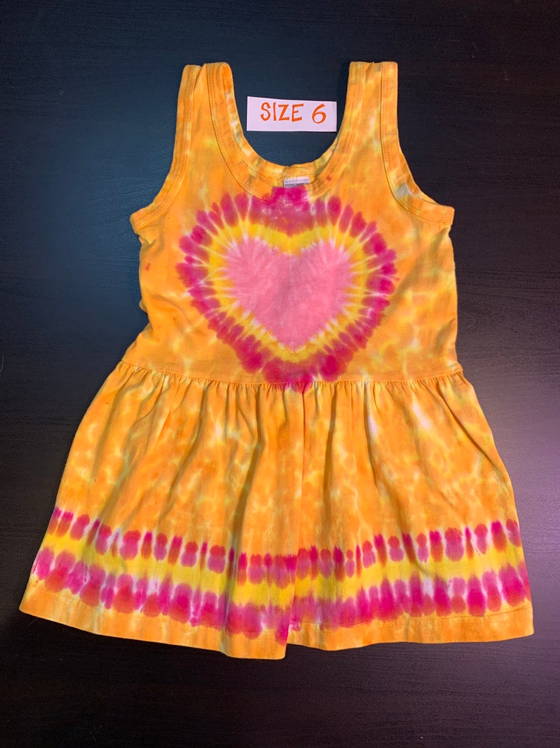 May include: A yellow, pink, and white tie-dye sleeveless dress with a heart design. The dress has a ruffled hem and says "SIZE 6" on the tag.