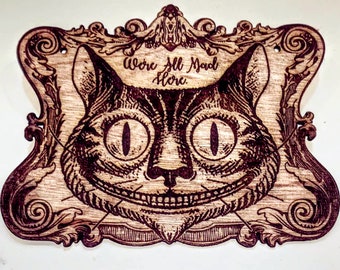 Könnte beinhalten: Eine lasergravierte Holzplatte mit dem Gesicht einer grinsenden Katze mit großen Augen und Schnurrhaaren. Das Design ist von einem verzierten Rahmen mit Schnörkeln umgeben. Über der Katze steht der Text: "We're All Mad Here". Das Holz ist naturbelassen.