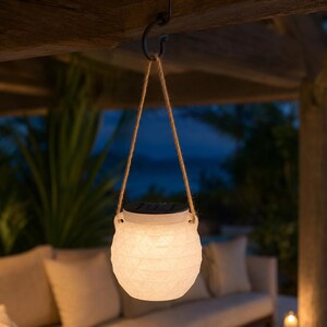 May include: A white, textured, spherical solar lantern with a geometric pattern, suspended by a tan jute rope from a metal hook. The lantern features a black solar panel on its top surface and emits a soft, warm glow from within its body.