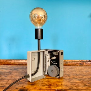 Peut inclure: Une lampe de table de style vintage réalisée à partir d'un projecteur de film reconditionné. La lampe a une base en métal gris avec un cordon noir et une ampoule en verre clair avec une lueur chaude. Le mécanisme du projecteur est visible sur le côté de la base.