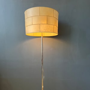 May include: A chrome floor lamp with a white fabric shade. The shade has a brick pattern and is lit with a warm light bulb.
