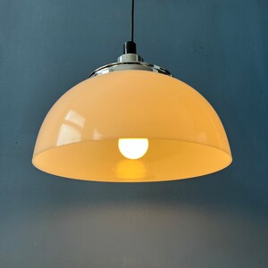 May include: A white, dome-shaped pendant light fixture with a chrome trim hangs from a black cord against a blue wall. The light is on and illuminates the room.