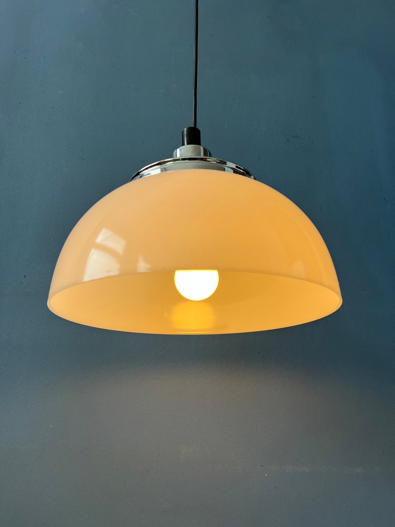 May include: A white, dome-shaped pendant light fixture with a chrome trim hangs from a black cord against a blue wall. The light is on and illuminates the room.