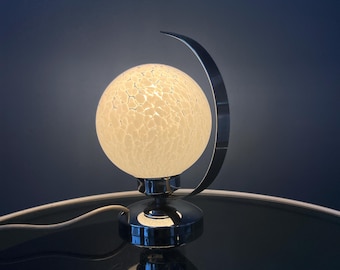 May include: A chrome table lamp with a white globe-shaped shade. The shade has a textured, crackled glass finish. The lamp base is a crescent shape.