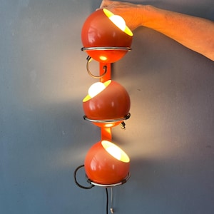 May include: A wall-mounted lamp featuring three spherical orange shades arranged vertically on a central orange bar. Each sphere is held by a chrome-finished metal ring and emits a warm light from an internal bulb. A black power cord hangs below.