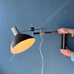 May include: A wall-mounted lamp with a dark brown conical shade and a chrome arm. The lamp is attached to a dark wooden base. The cord is white. The lamp is lit, casting a warm glow.