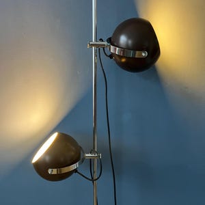 Puede incluir: Una lámpara de pie vintage con dos luminarias esféricas ajustables de color marrón oscuro. Cada luz tiene una banda plateada y está montada en un poste cromado. Las luces están orientadas para iluminar el área.