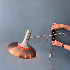 Könnte beinhalten: Eine Wandleuchte mit einem breiten, halbtransparenten, kupferfarbenen Glasschirm und einem Hals aus gebürstetem silberfarbenem Metall. Die Leuchte verfügt über einen verstellbaren, mehrfach gegliederten Chromarm an einem dunklen Holzsockel.