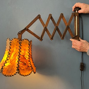 Może przedstawiać: Drewniana lampa ścienna z składanym ramieniem i brązowym abażurem ze skóry. Lampa jest włączona, a abażur jest oświetlony.