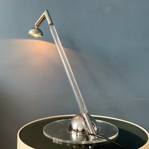 May include: A chrome desk lamp with a long, clear acrylic arm and a round, chrome base. The lamp is on and casting a warm light.