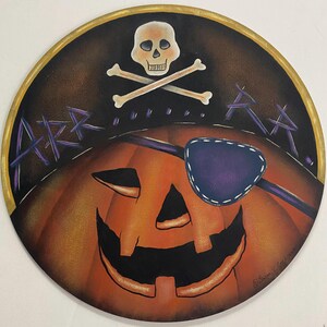 May include: A circular wall plaque featuring a painted orange jack-o'-lantern with a black eye patch, a skull and crossbones symbol above it, and the purple text "ARR.....RR." set against a dark background with a gold-painted circular border.