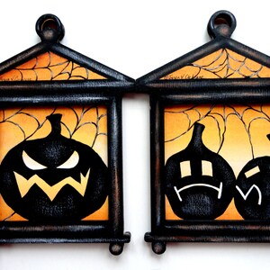 Puede incluir: Dos cuadros enmarcados en madera negra con fondos naranja y negro. Las imágenes presentan siluetas negras de calabazas con diferentes expresiones faciales. La imagen superior presenta una calabaza con una expresión de enfado. La imagen inferior presenta dos calabazas con expresiones tristes.