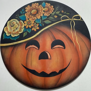 May include: A circular wooden plaque featuring a hand-painted orange jack-o'-lantern with a smiling face. The pumpkin wears a black hat adorned with yellow and orange flowers, green leaves, and a thin gold ribbon tied in a bow on the side.
