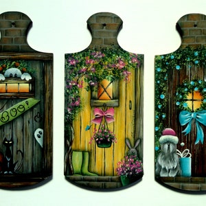 May include: Three wooden door-shaped signs with painted scenes. The first sign has a black cat, a pumpkin, and a ghost with the text "BOO!" on it. The second sign has a yellow door with a pink bow and a hanging flower pot. The third sign has a brown door with a blue bow and a dog wearing a Santa hat.