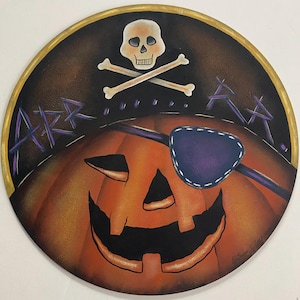 May include: A circular wall plaque featuring a painted orange jack-o'-lantern wearing a purple eye patch. Above the pumpkin is a white skull and crossbones on a black background with the purple text ARR......RR. The plaque has a gold rim.