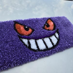 May include: Purple rug with a white and black smiling face and orange eyes. The rug is made of a soft, fuzzy material.