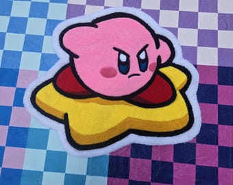 May include: A tufted rug shaped like the character Kirby, featuring a round pink body with dark red feet and a determined expression, riding on a yellow star. The entire design is outlined in black and bordered by a thick white tufted edge.