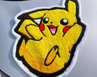 Può includere: Un tappeto giallo con il personaggio di Pikachu con contorni neri e cuciture bianche. Il tappeto è realizzato in un materiale morbido e peluche e presenta un Pikachu sorridente con le guance rosse.