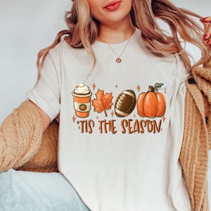 K&ouml;nnte beinhalten: Wei&szlig;es T-Shirt mit einem Aufdruck einer K&uuml;rbis, einem Football, einem Ahornblatt und einer Tasse Kaffee mit Schlagsahne. Der Text "'Tis the season" befindet sich unter dem Aufdruck.