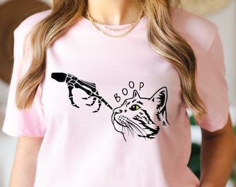 May include: Pink t-shirt featuring a black and white graphic of a skeleton hand reaching out to boop a cat's nose. The text "BOOP" is above the cat.