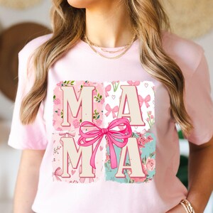 May include: A light pink t-shirt featuring the word "MAMA" in a floral design. The letters are in a light beige color, set against four squares with pink floral patterns, bows, and hearts. A large pink bow is centered over the letters.