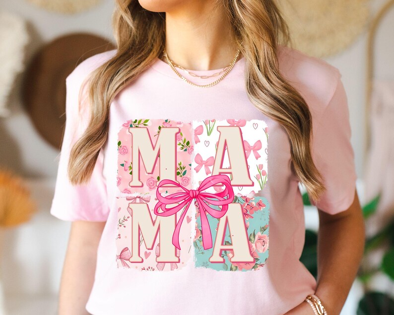 May include: A light pink t-shirt featuring the word "MAMA" in a floral design. The letters are in a light beige color, set against four squares with pink floral patterns, bows, and hearts. A large pink bow is centered over the letters.