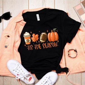 K&ouml;nnte beinhalten: Schwarzes T-Shirt mit einem Motiv aus einer Kaffeetasse, einem Blatt, einem Football und einem K&uuml;rbis. Der Text "'Tis the season" befindet sich unter dem Motiv.