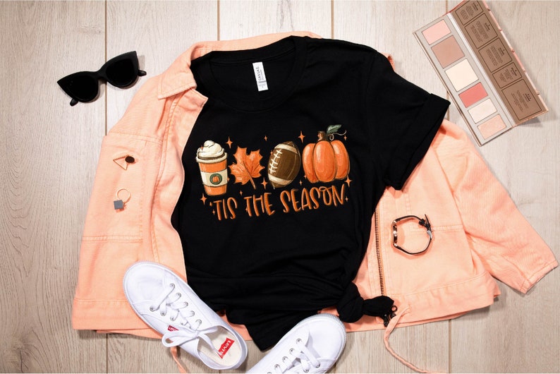 K&ouml;nnte beinhalten: Schwarzes T-Shirt mit einem Motiv aus einer Kaffeetasse, einem Blatt, einem Football und einem K&uuml;rbis. Der Text "'Tis the season" befindet sich unter dem Motiv.