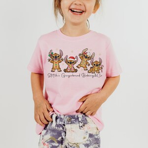 Puede incluir: Una camiseta rosa claro con un gr&aacute;fico de cuatro personajes Stitch estilo pan de jengibre con decoraciones navide&ntilde;as. El texto "Stitch's Gingerbread Baking & Co" est&aacute; impreso debajo. La camiseta la lleva un ni&ntilde;o peque&ntilde;o.