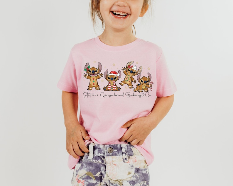 Puede incluir: Una camiseta rosa claro con un gr&aacute;fico de cuatro personajes Stitch estilo pan de jengibre con decoraciones navide&ntilde;as. El texto "Stitch's Gingerbread Baking & Co" est&aacute; impreso debajo. La camiseta la lleva un ni&ntilde;o peque&ntilde;o.