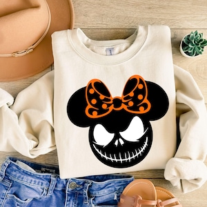 Puede incluir: Sudadera color crema con un gr&aacute;fico negro y naranja de una cara con un lazo. El gr&aacute;fico tiene una cara negra con una sonrisa y ojos blancos, y un lazo naranja con lunares negros. La sudadera se combina con vaqueros azules y sandalias.