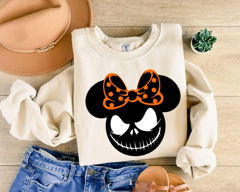 Puede incluir: Sudadera color crema con un gr&aacute;fico negro y naranja de una cara con un lazo. El gr&aacute;fico tiene una cara negra con una sonrisa y ojos blancos, y un lazo naranja con lunares negros. La sudadera se combina con vaqueros azules y sandalias.