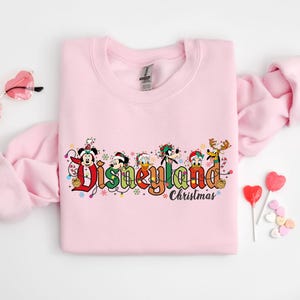 May include: A light pink sweatshirt featuring the text "Disneyland Christmas" in colorful lettering with Disney characters. The design includes Mickey Mouse, Donald Duck, Goofy, and other characters in festive holiday attire.