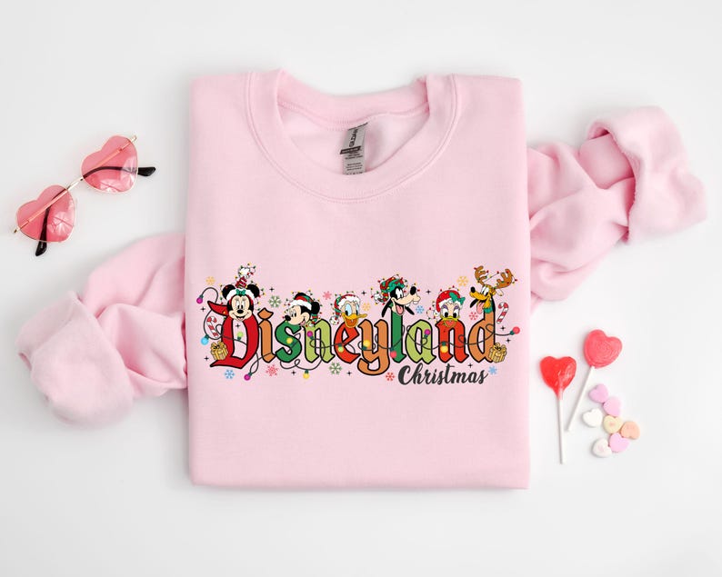 May include: A light pink sweatshirt featuring the text "Disneyland Christmas" in colorful lettering with Disney characters. The design includes Mickey Mouse, Donald Duck, Goofy, and other characters in festive holiday attire.