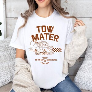 May include: White t-shirt featuring a brown graphic of Tow Mater from the movie Cars. The design includes the text "TOW MATER" above an illustration of the character, with "Piston Cup Racing Series 5 Time Champ" below.