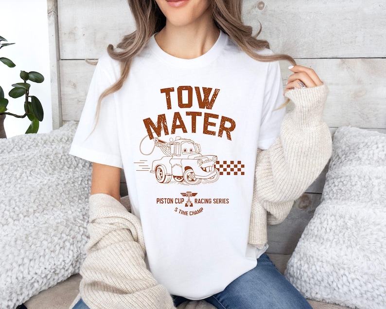 May include: White t-shirt featuring a brown graphic of Tow Mater from the movie Cars. The design includes the text "TOW MATER" above an illustration of the character, with "Piston Cup Racing Series 5 Time Champ" below.