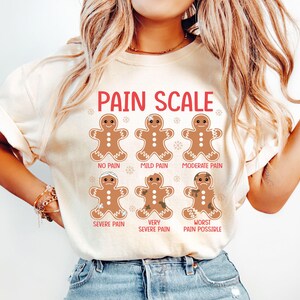 Op de afbeelding: Crèmekleurig T-shirt met een rode "PAIN SCALE" afbeelding. De afbeelding toont peperkoekmannetjes die pijnniveaus illustreren, van "No Pain" tot "Worst Pain Possible". Het shirt is een casual, comfortabele stijl.