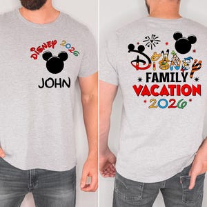 May include: A light gray t-shirt with a colorful Disney design. The front of the shirt features the text "Disney 2026" above a black Mickey Mouse head and the name "JOHN". The back of the shirt has "Disney Family Vacation 2026" with character graphics.